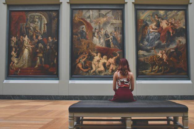 A young woman with dark hair sits on a bench in an art gallery. She is facing away from the camera toward three large paintings on the wall.