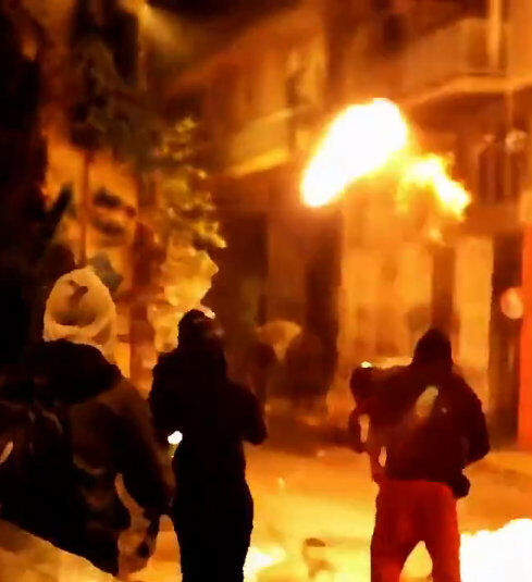 A chaotic scene of revolution tinted in yellow. In the foreground are three individuals dressed in dark hoodies fight dark mojoe on their revolution for life mastery with Spin Positive. They are facing away from the camera. The one on the far right throws a lit molatov cocktail in the direction they are facing. In the background are buildings. The streets are littered with debris.
