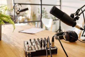 A wooden desk supports two podcast host stations, each with its own broadcast microphone on a boom arm mount. One in the background and another in the foreground. On the left hand side is a fern-like plant. In the foreground is a two-channel mixer with four cables running from it. To the right is a pair of studio headphones. The background station has a notepad and pen. In the background is a window allowing in soft natural light. Outside of the window some plants live.