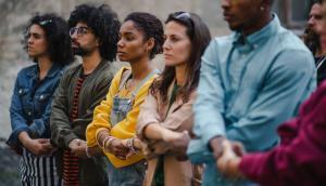 A group of people hold hands with crossed arms. The first is an olive skinned lady with a denim jacket. Next to here is an olive-skinned man with a luscious hair of dark curly locks. To his left is a black woman wearing a brightly colored sweatshirt and blue overalls. To her left is a white woman with long dark hair, sunglasses on her head and a neutral colored jacket. The final person in the line that we can see is a black man. He is tall and wearing a light blue, button up shirt. All appear to be forming a line of solidarity at a protest of some ilk.